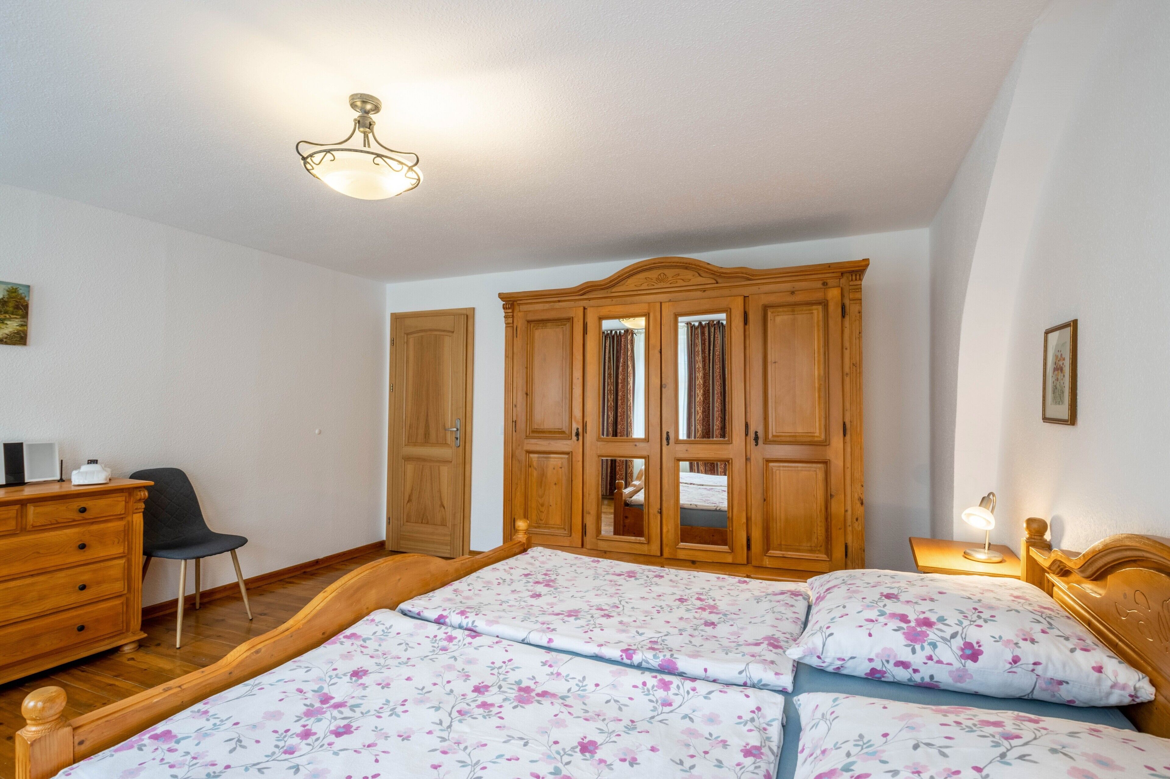 A bedroom with a wooden wardrobe, a bed with floral bedding, a desk with a chair, and a ceiling light fixture.