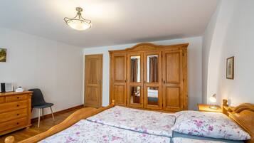 A bedroom with a wooden wardrobe, a bed with floral bedding, a desk with a chair, and a ceiling light fixture.