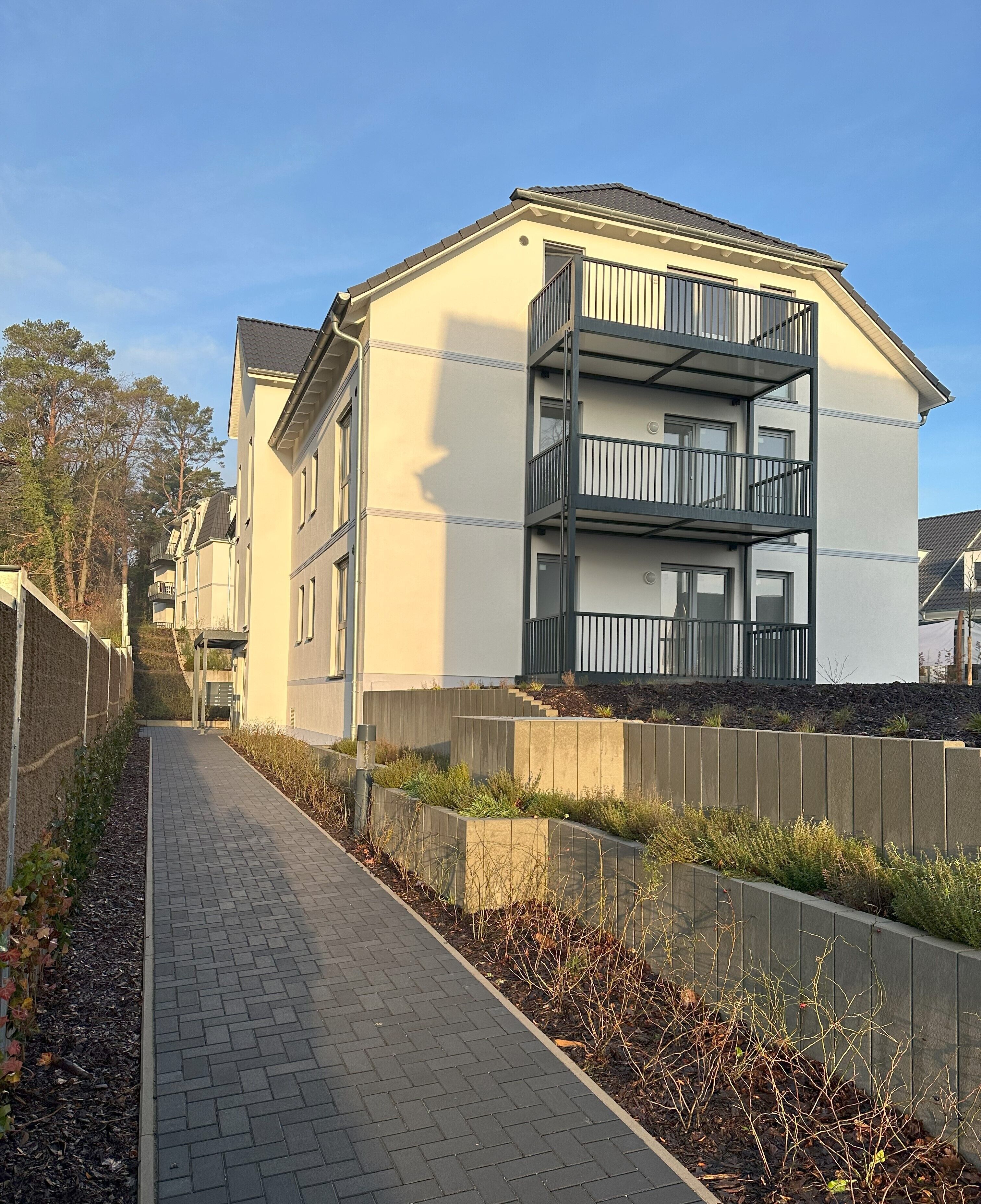 A multi-story residential building with balconies, a paved walkway, and a landscaped area with plants.