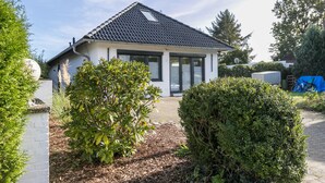 Einbungalow mit Ziegeldach, einer überdachten Veranda und einem Garten mit Sträuchern und einem Baum.