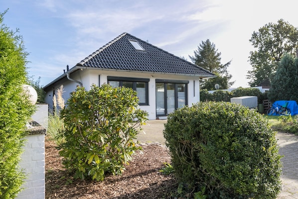 Une maison de plain-pied avec un toit en tuiles, une véranda et un jardin agrémenté d’arbustes et d’un arbre.