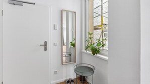 Een witte deur met een metalen handvat, een klein tafeltje met een metalen frame, een spiegel en een raam met uitzicht op een gebouw.
