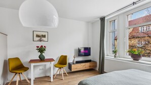 Een moderne slaapkamer met een bed, een tv waarop 'NETFLIX' staat, twee stoelen, een klein tafeltje, een vaas met bloemen en een raam met uitzicht op gebouwen.