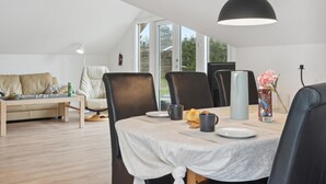 Un coin repas avec une table dressée pour deux personnes, des chaises, un luminaire suspendu et une vue sur l’extérieur grâce à de grandes fenêtres.