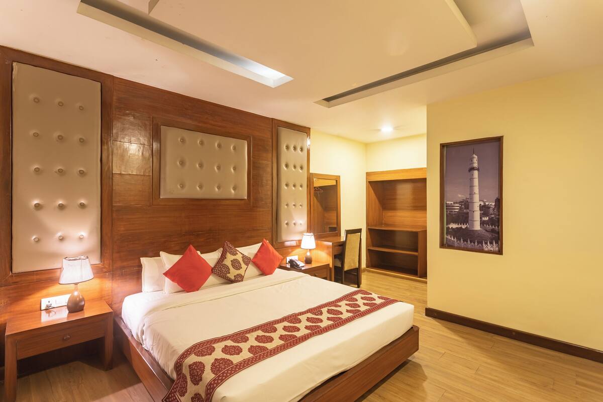 A hotel room with a large bed, wooden headboard, a nightstand with a lamp, a chair, a wardrobe, and a framed picture on the wall.
