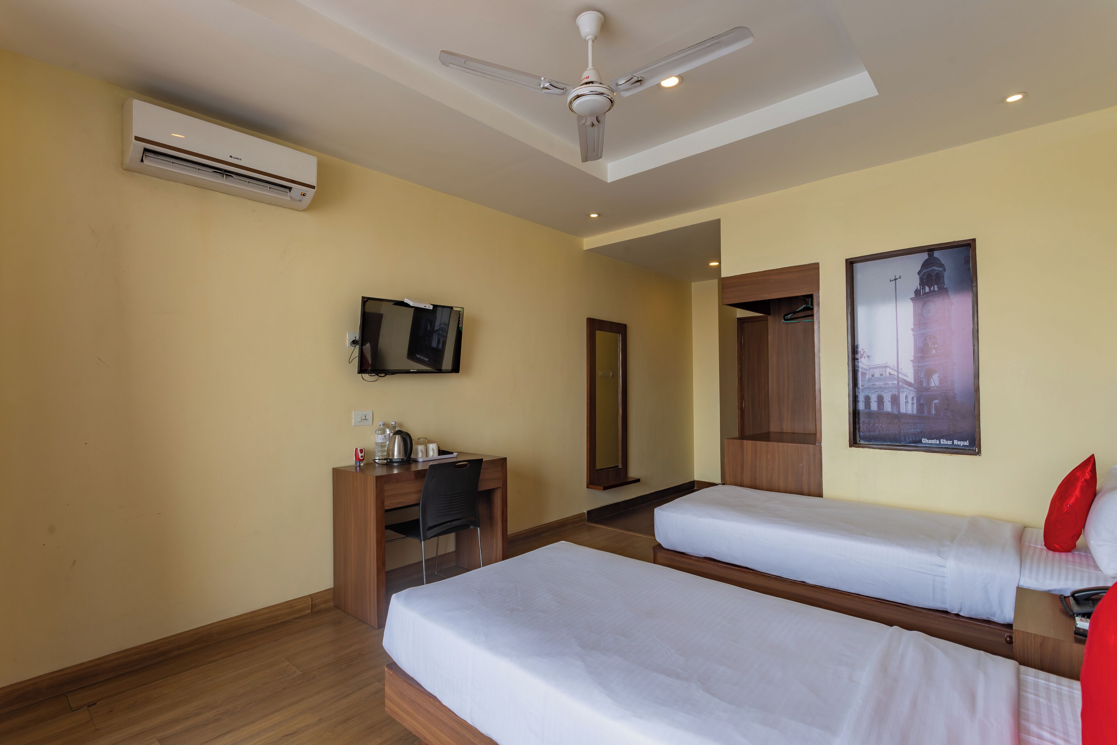 A hotel room with two single beds, a desk with a chair, an air conditioner, a ceiling fan, a TV, and a poster on the wall.