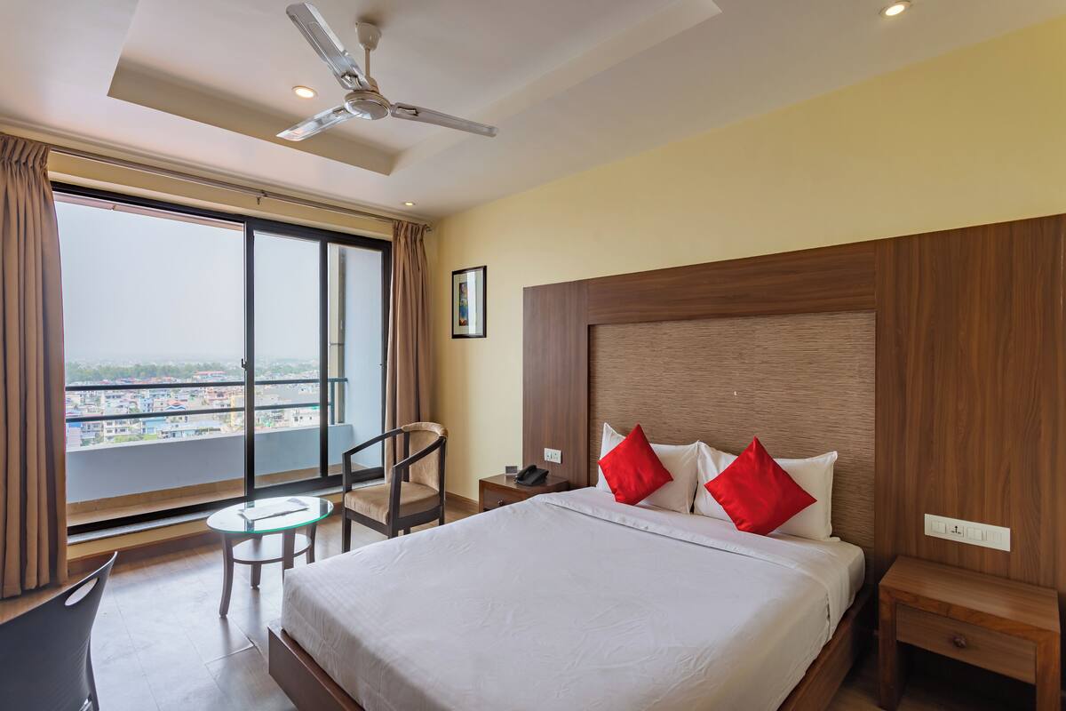 A hotel room with a large bed, a desk, a chair, a small table, a nightstand, and a ceiling fan.