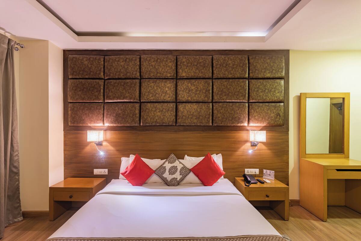 A hotel room with a large bed, two bedside tables, a wooden headboard, a mirror, and a nightstand with a phone and a lamp.