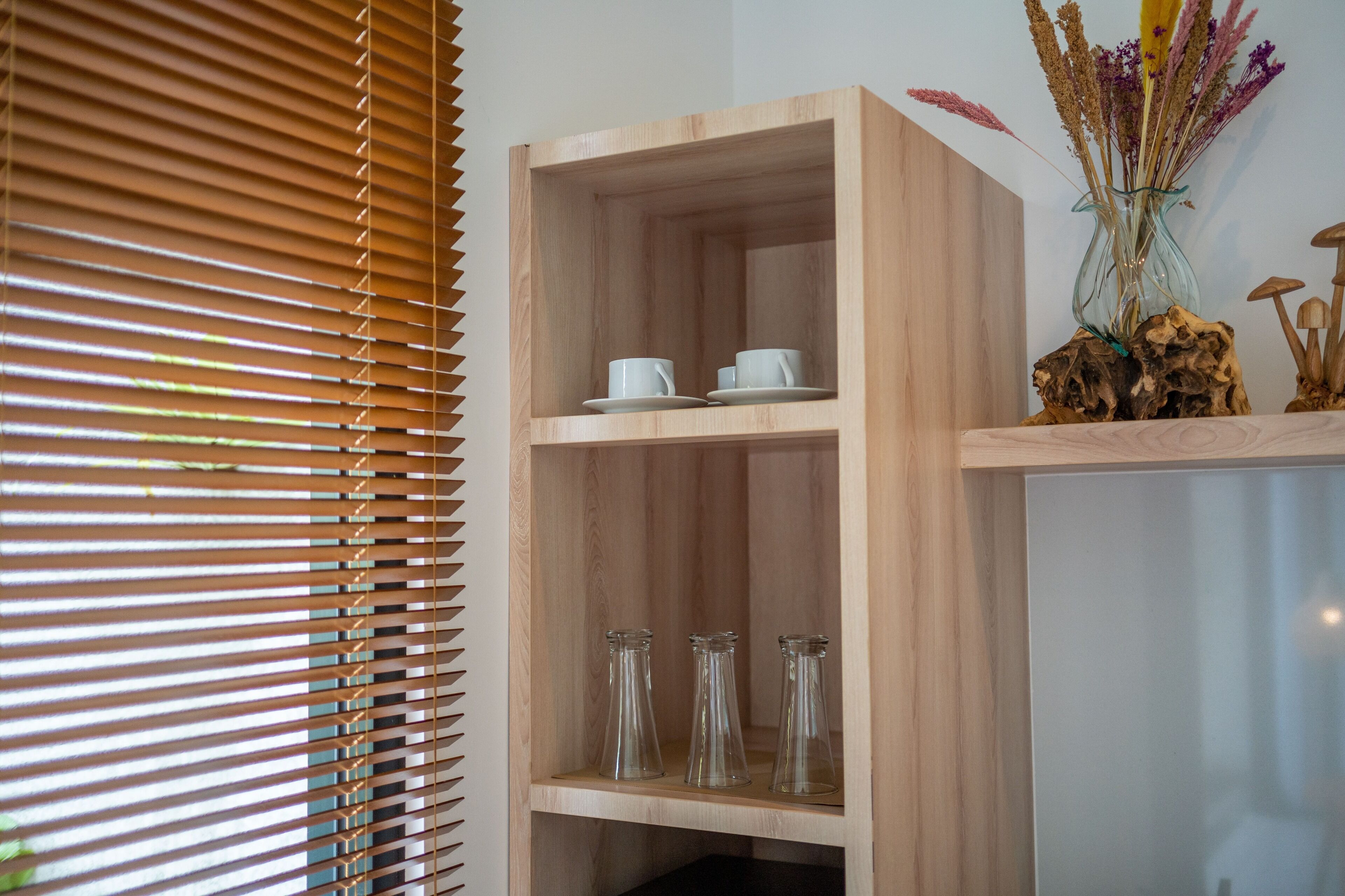 A wooden shelf with white cups and glass bottles, a vase with dried flowers, and a wooden sculpture.