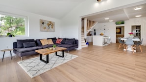 Un salon moderne avec un canapé gris foncé, une table basse en bois et un tapis blanc. On aperçoit à l’arrière-plan un coin repas avec une table et des chaises blanches.