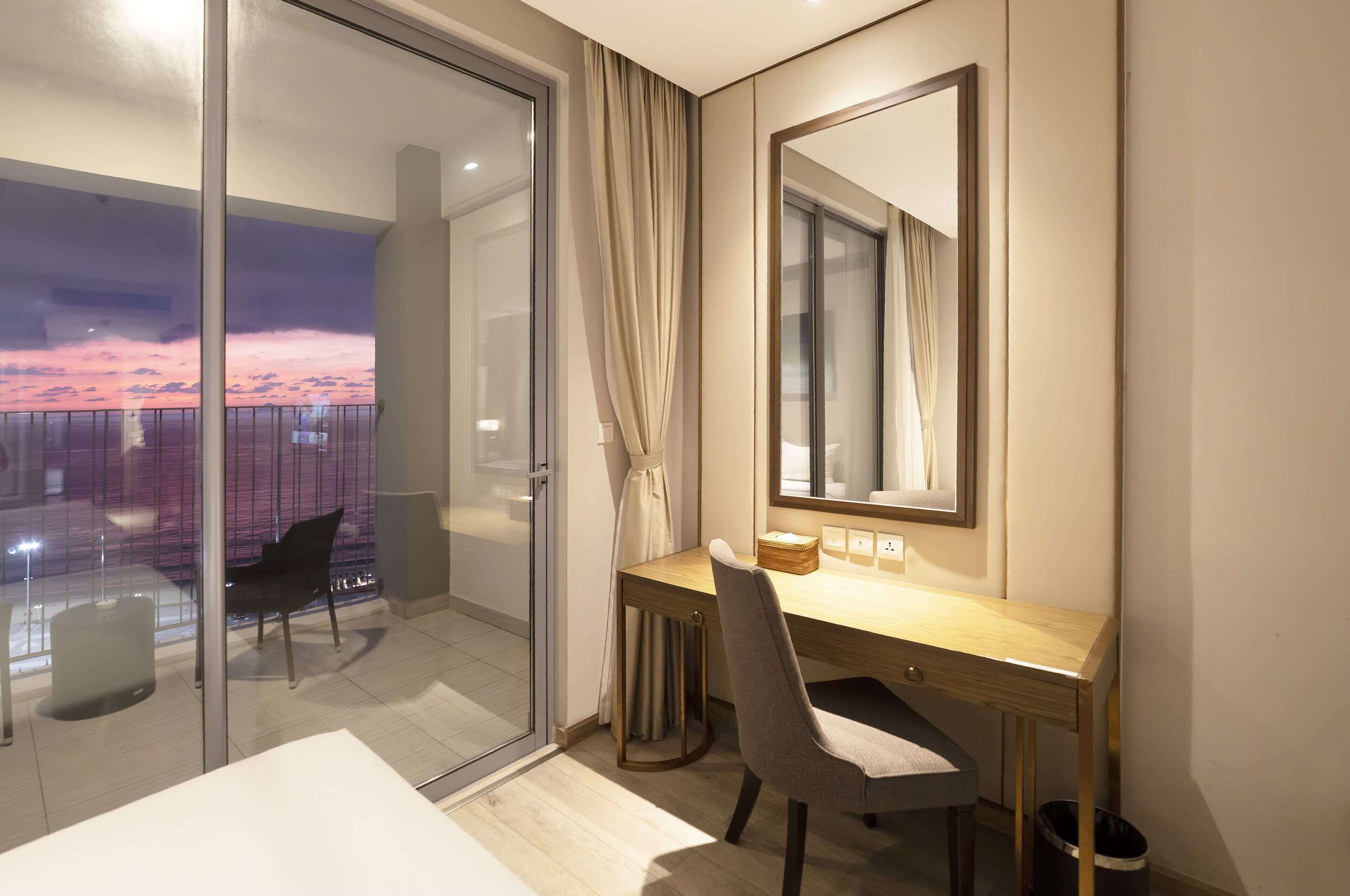 A modern hotel room with a large window, a desk with a chair, a bedside table, and a mirror.