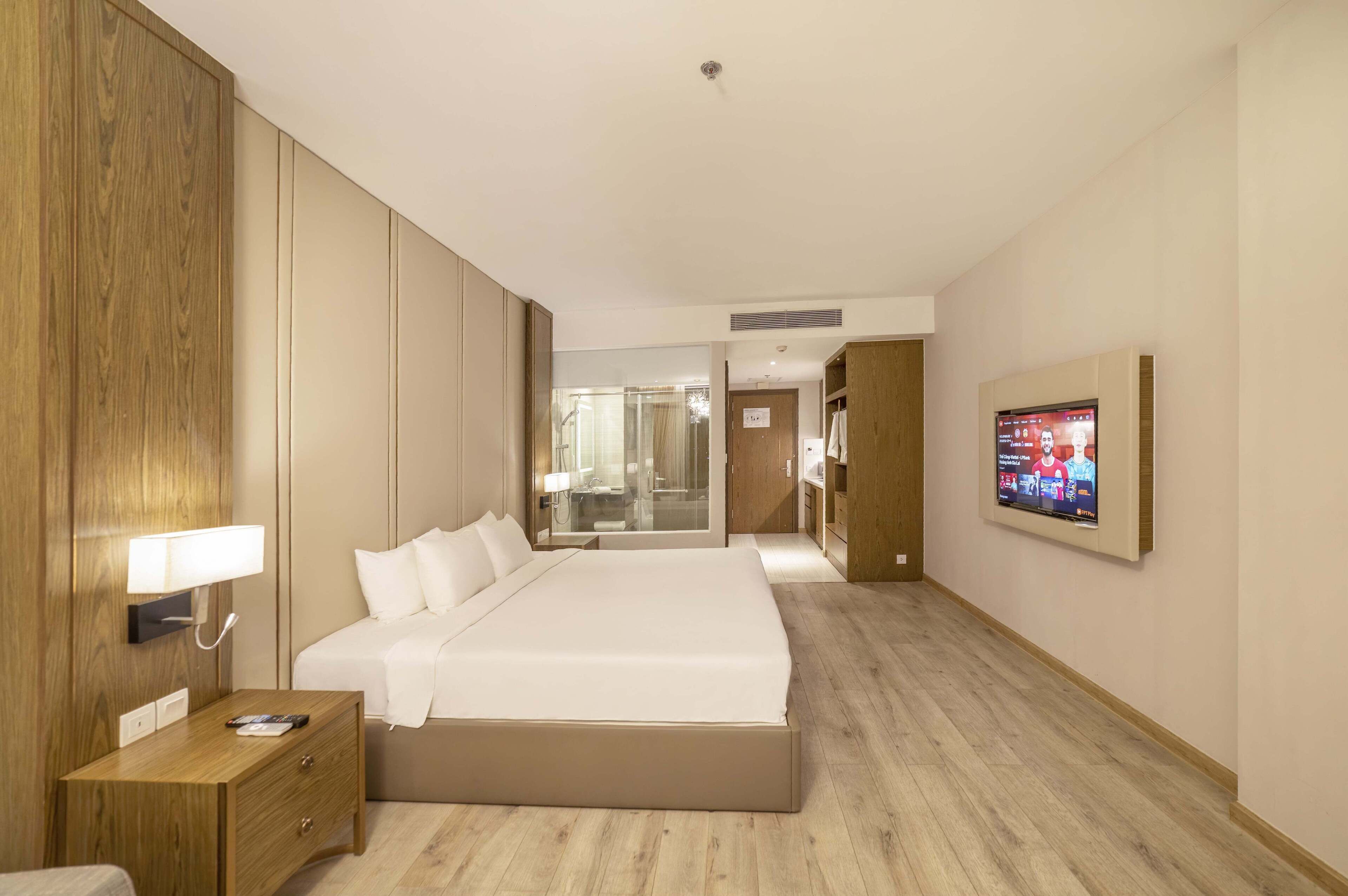 A hotel room with a large bed, a bedside table with a lamp, a TV mounted on the wall, and a wooden wardrobe.