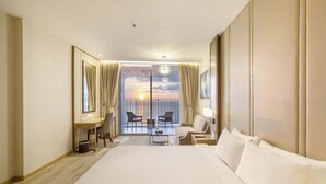 A hotel room with a large bed, a desk, a chair, a small table, and a balcony with a view of the sunset.