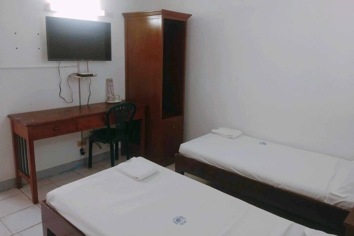 Una habitació amb escriptori de fusta, una cadira, un llit i televisió penjada a la paret.