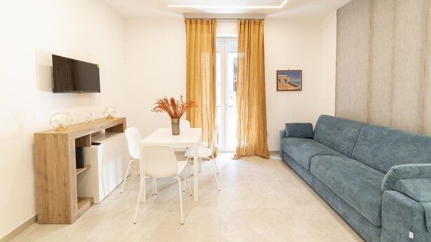 A modern living room with a sofa, a dining table with chairs, a TV mounted on the wall, and a window with curtains. - Vico Cangiani al Mercato 20 (Naples)