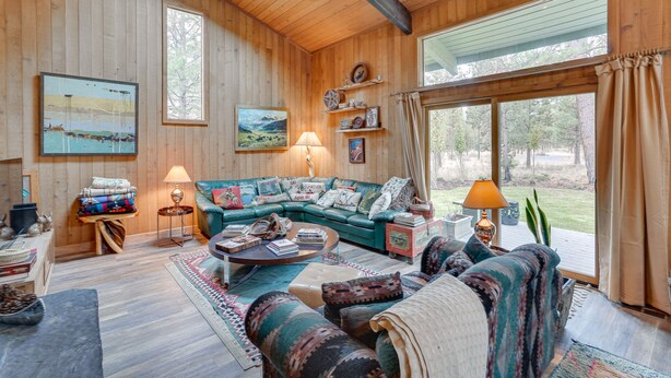 A cozy cabin interior with a large sofa, a coffee table, and a fireplace. There are wooden walls and a large window offering a view of the outdoors. - Sunriver Resort Home: Near Skiing, Town & Trails (Sunriver)
