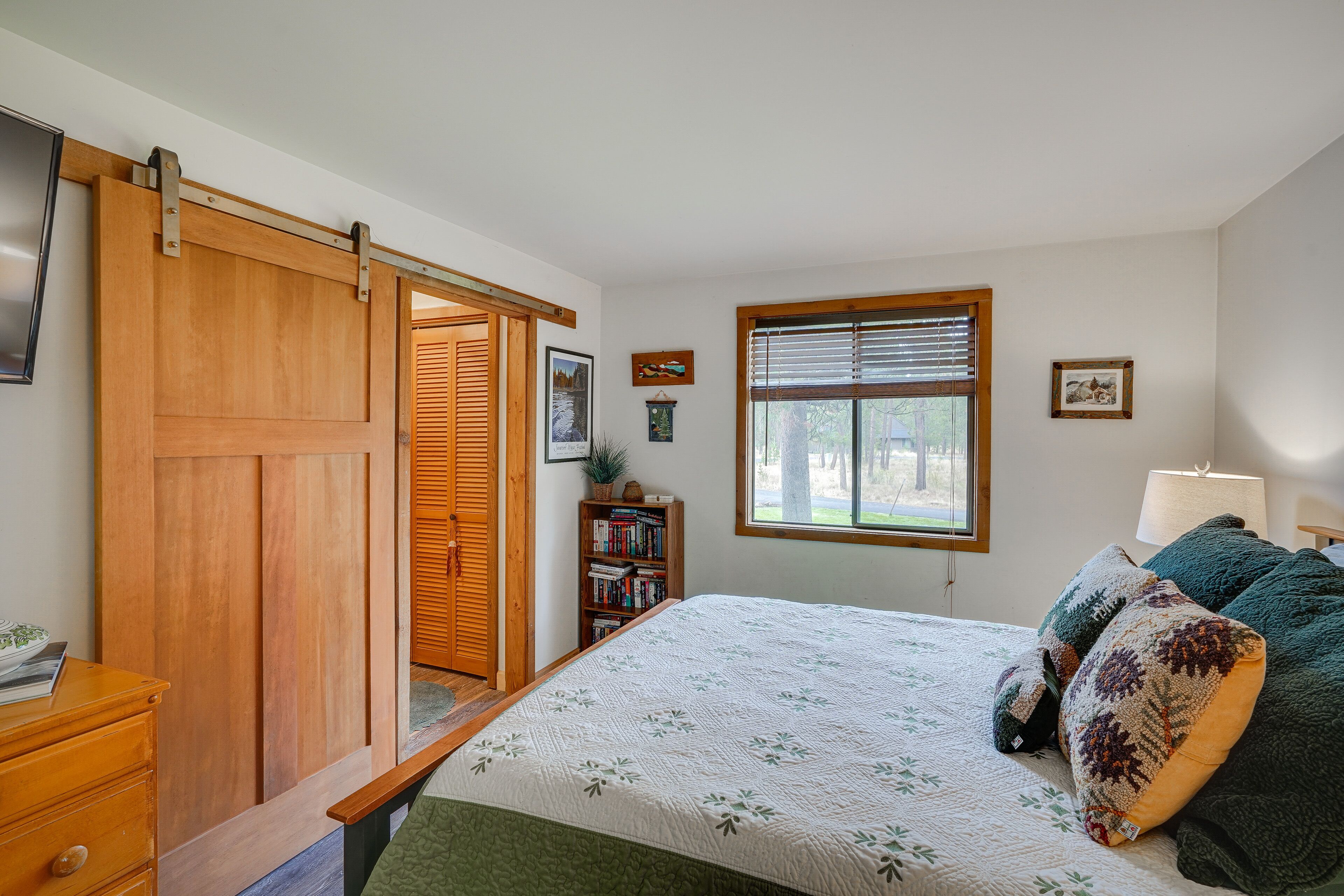 A bedroom with a bed, a wooden wardrobe, a bookshelf, a window with blinds, and a lamp.