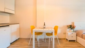 A compact kitchen with a white refrigerator, a small dining table with two chairs, and a bedside table with a lamp and a flower vase. - Mono Garden by Interhome (Marina di Bibbona)