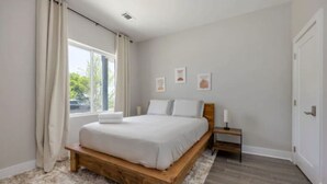 A bedroom with a wooden bed, a small bedside table, a window with curtains, and a door. - Luxury Home w/ Rooftop & 8 Beds! 10 to Downtown! (Nashville)