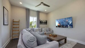 A modern living room with a grey sectional sofa, a coffee table, and a mounted TV displaying a concert. - Luxury Home w/ Rooftop & 8 Beds! 10 to Downtown! (Nashville)
