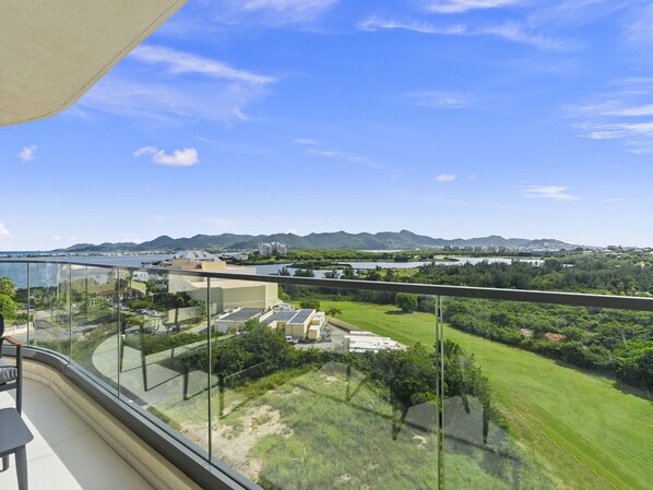 A balcony with a clear view of a coastal landscape, including a beach, mountains, and a body of water. - B-904 Panoramic ocean view 1 bedroom cond (Cupecoy)
