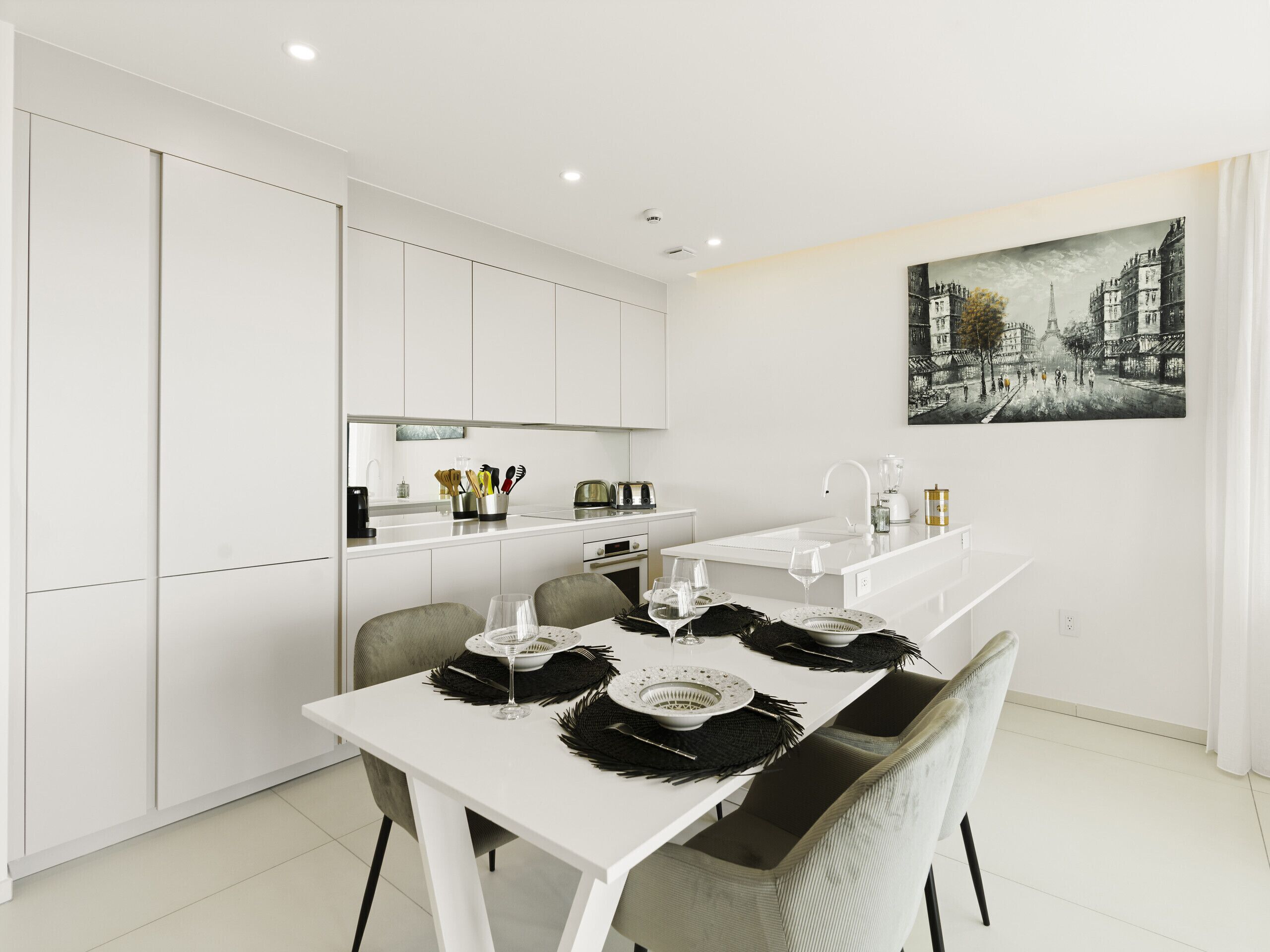 A modern kitchen with white cabinetry, a dining table set with black placemats and white plates, and a painting of a cityscape on the wall.