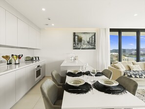A modern kitchen with white cabinets, a dining area with a white table set for two, and a large window offering a city view. - B-904 Panoramic ocean view 1 bedroom cond (Cupecoy)