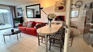 A modern living room with a red sofa, a dining table with chairs, a coffee table, and a vase of flowers. There is a large mirror on the wall, a clock, and a view of the outside through a sliding door.