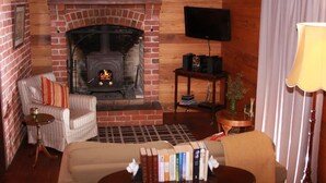 Un salon chaleureux avec une cheminée en briques, un téléviseur fixé au mur, un fauteuil à rayures, une petite table avec une lampe et une bibliothèque.