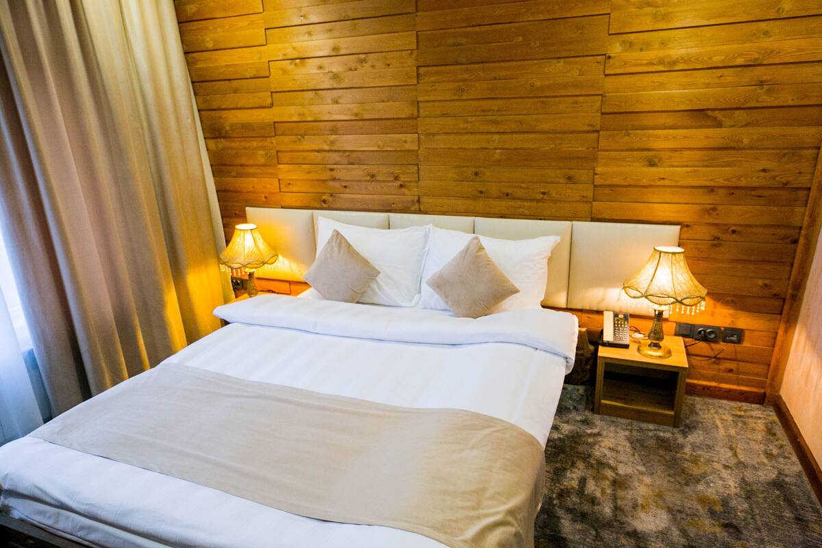 Een hotelkamer met een houten hoofdeinde, een bed met witte lakens, twee bedlampjes en een vloerbedekking.