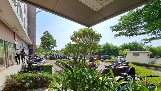 A covered parking area with motorcycles and scooters parked, surrounded by greenery and a building with glass doors. - OYO Life 93140 Apartment Grand Sentraland By Fahri (Karawang)
