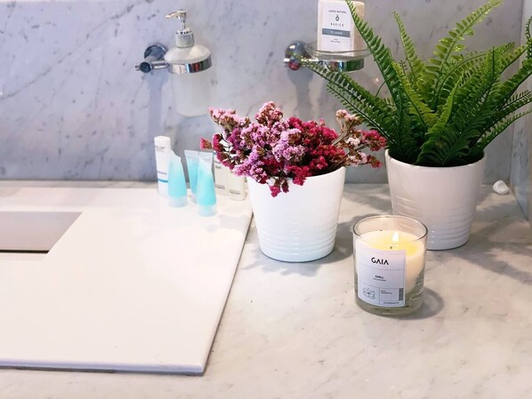 Una encimera de baño con un lavabo blanco, una planta en maceta, una vela con la etiqueta "GAIA CHILL" y varios productos para el cuidado de la piel.