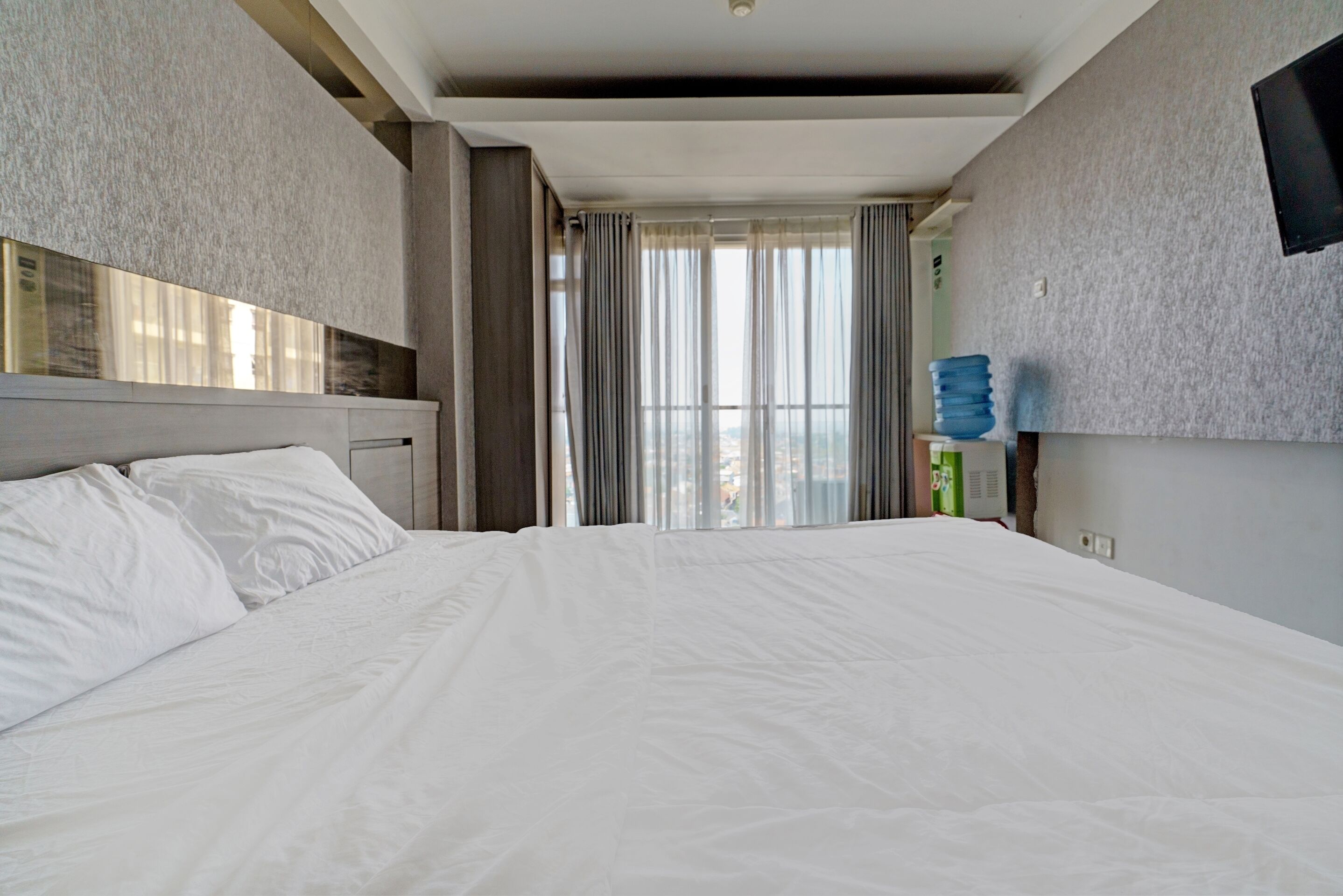 A hotel room with a large bed, a headboard, a flat-screen TV mounted on the wall, and a window with curtains.