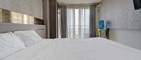 A hotel room with a large bed, a headboard, a flat-screen TV mounted on the wall, and a window with curtains.
