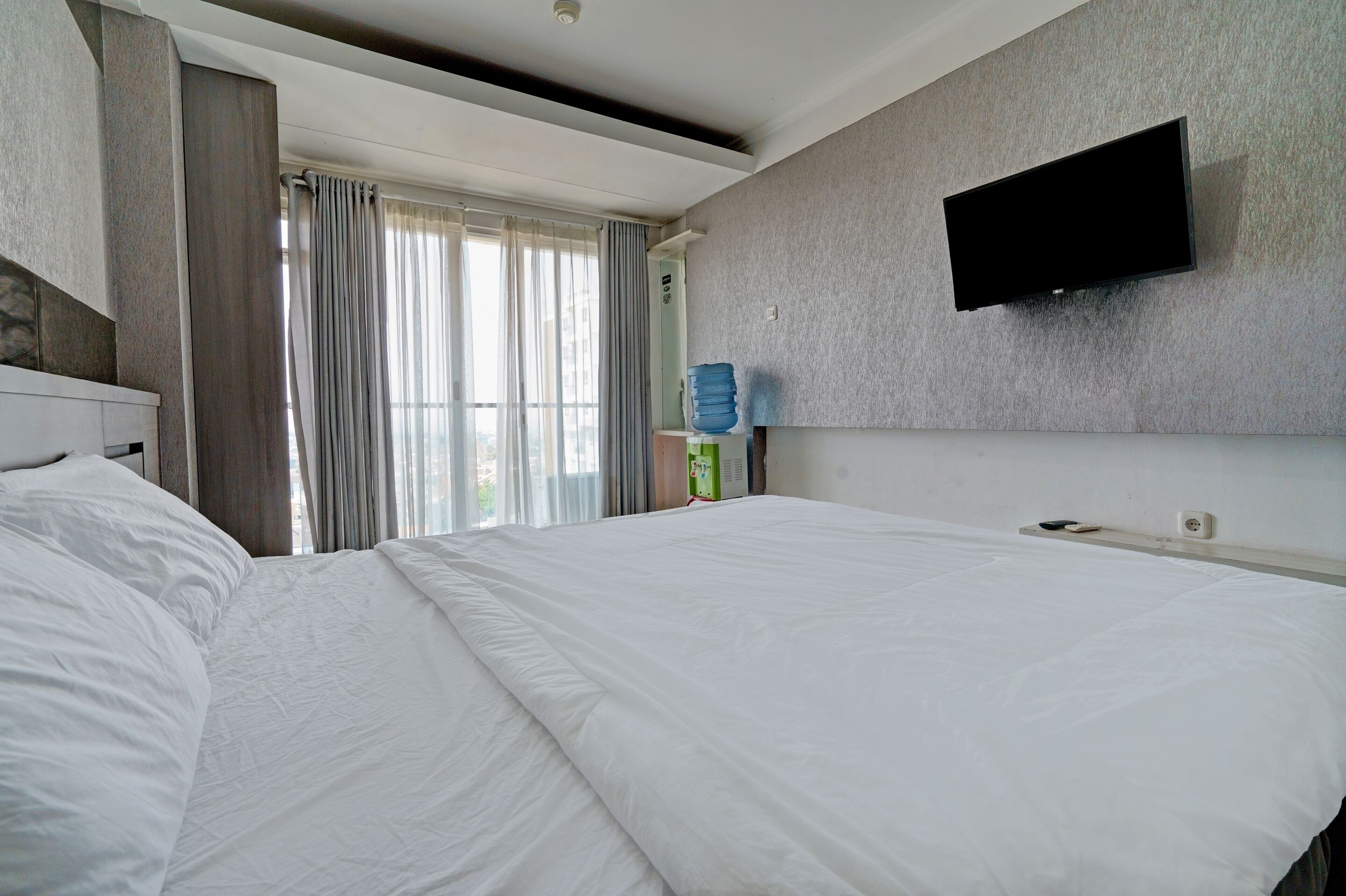 Habitación de hotel con una cama grande, un televisor de pantalla plana en la pared, un dispensador de agua y una ventana con cortinas.