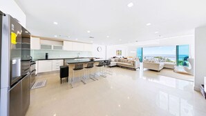Una cocina moderna con una isla central, un comedor con taburetes de bar, una sala de estar con sofá y vista al mar, y un ventanal grande.