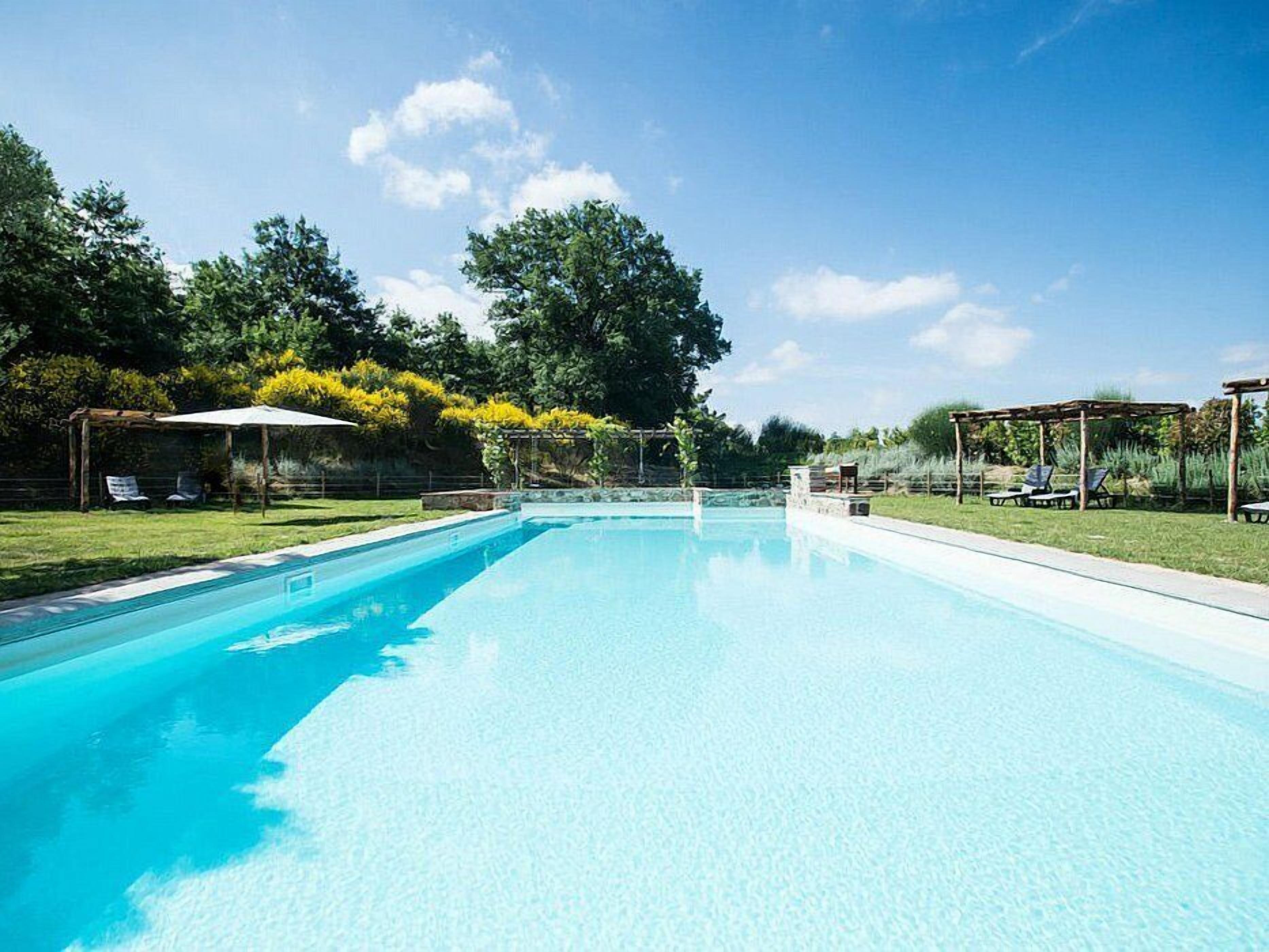 A rectangular swimming pool with clear blue water, surrounded by a grassy area with trees and a pergola.