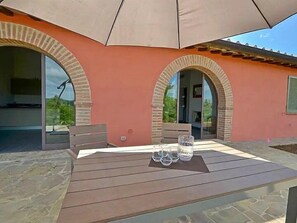 Una veranda esterna con tavolo di legno, due ingressi ad arco e un grande ombrellone.