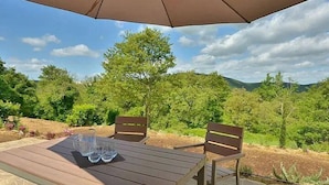 Une terrasse extérieure avec une table et des chaises, protégée par un grand parasol, et entourée d’une végétation luxuriante et de collines.