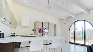 Une cuisine moderne dotée d’un îlot central, de tabourets de bar blancs et d’une grande fenêtre offrant une vue sur l’extérieur.