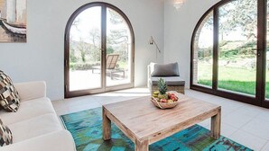 A spacious living room with a white sofa, a wooden coffee table with fruit, and large arched windows offering a view of the outdoors.