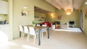 Ein geräumiger Wohnbereich mit Esstisch, Stühlen und Kamin. Eine Küchenzeile mit Schränken und Arbeitsplatte ist ebenfalls vorhanden. Der Raum verfügt über eine hohe Decke mit freiliegenden Balken und großen Fenstern für natürliches Licht.