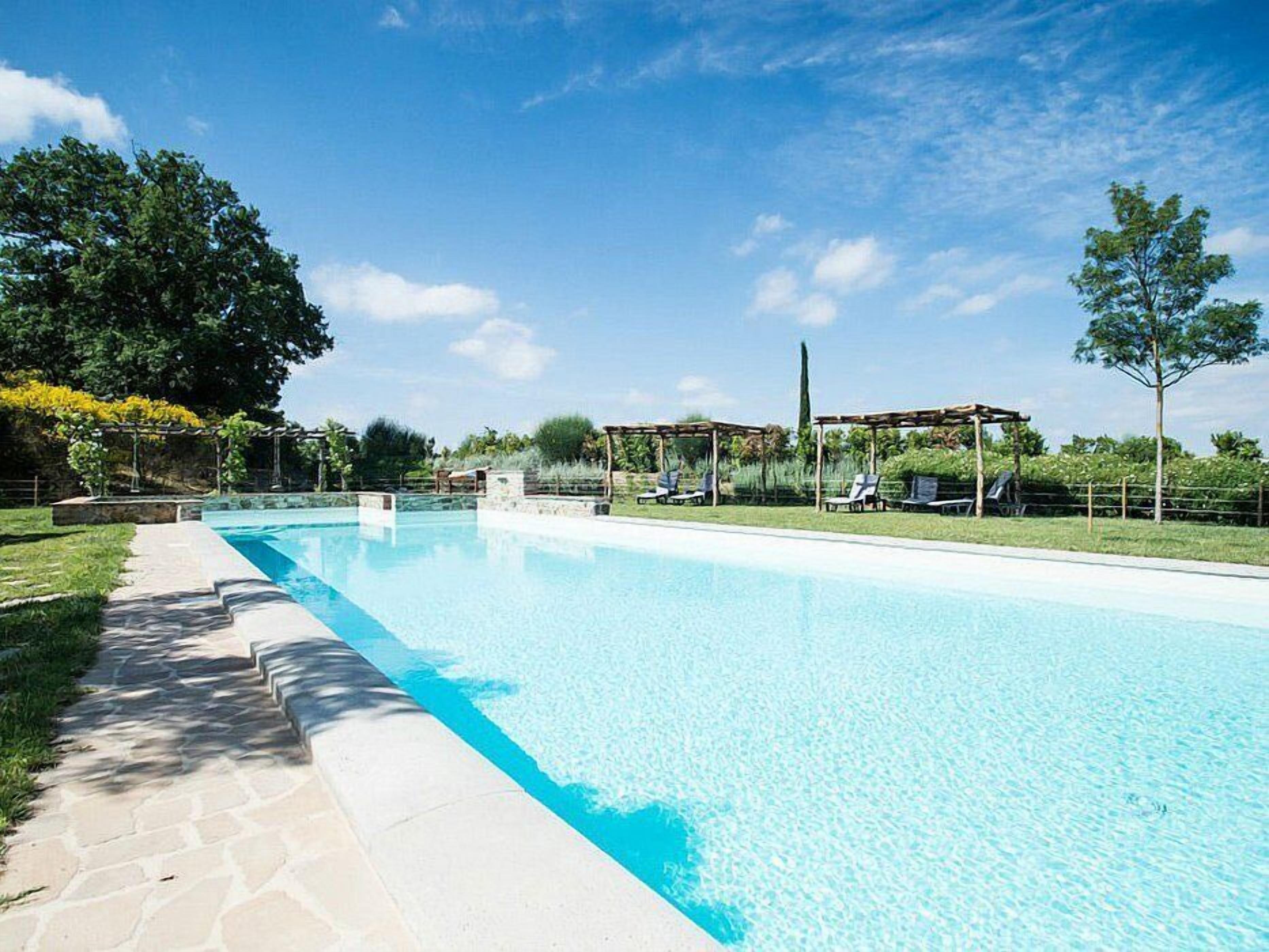 A rectangular swimming pool with clear blue water, surrounded by a stone pathway and shaded by pergolas.