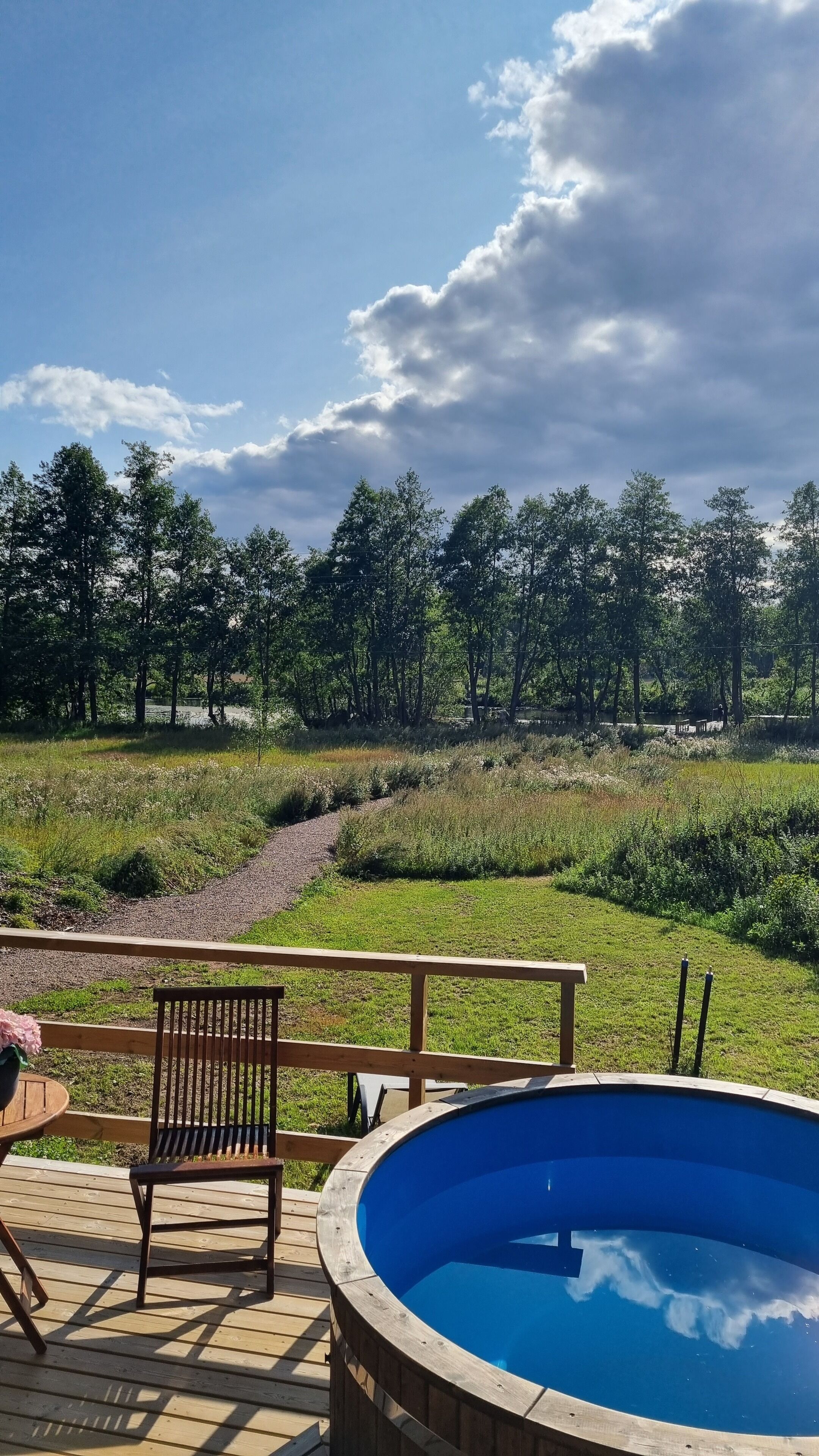 Una terraza de madera con piscina, una silla y vista a un área verde con árboles.