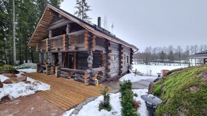 A log cabin with a wooden deck, surrounded by snow and trees.