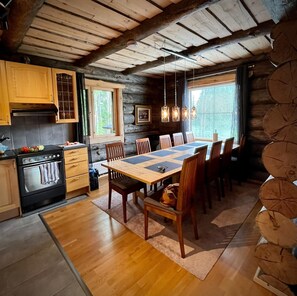 A rustic cabin interior with a wooden dining table, chairs, a kitchen with a stove, and a large window.