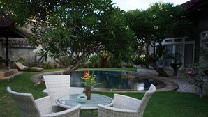 Mesa redonda con sillas de mimbre en un jardín con un estanque y vegetación.