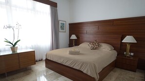Habitación de hotel con una cama grande, cabecera de madera, dos mesitas de noche con lámparas, un aparador y una orquídea en maceta, además de un cuadro en la pared.