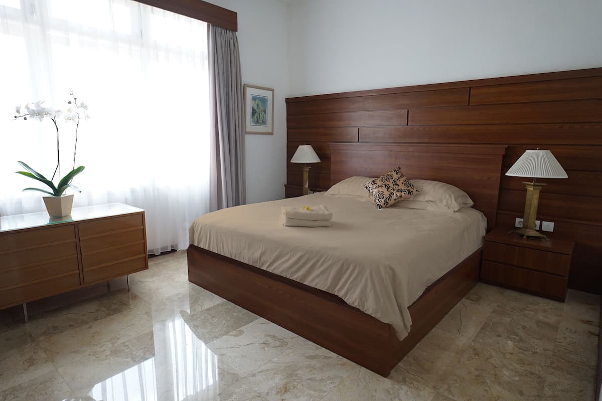 A hotel room with a large bed, wooden headboard, two bedside tables with lamps, a nightstand, a potted orchid, and a framed picture on the wall.