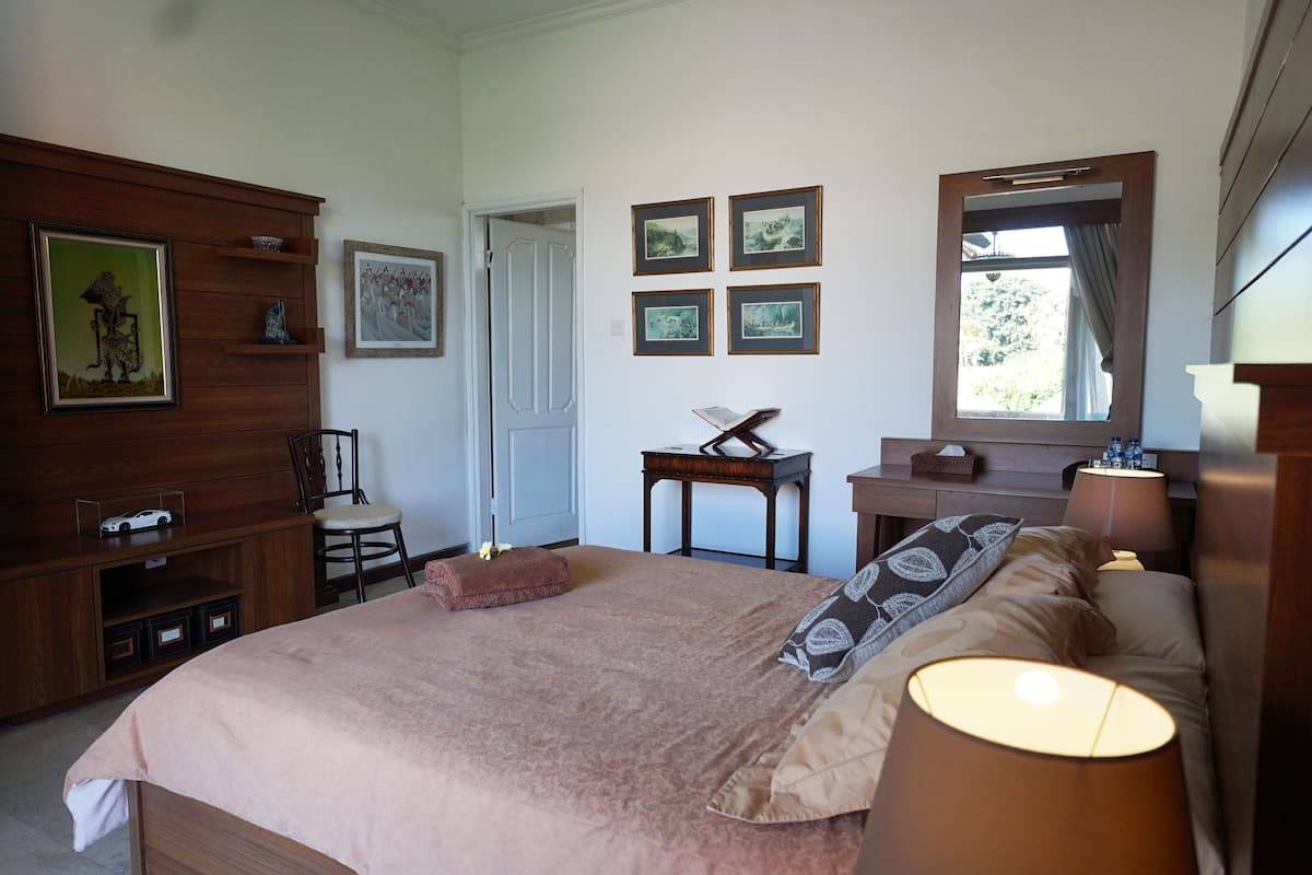A bedroom with a large bed, bedside lamps, a wooden dresser, a chair, and a small table with a lamp. There are framed pictures on the wall and a window with a view of greenery.